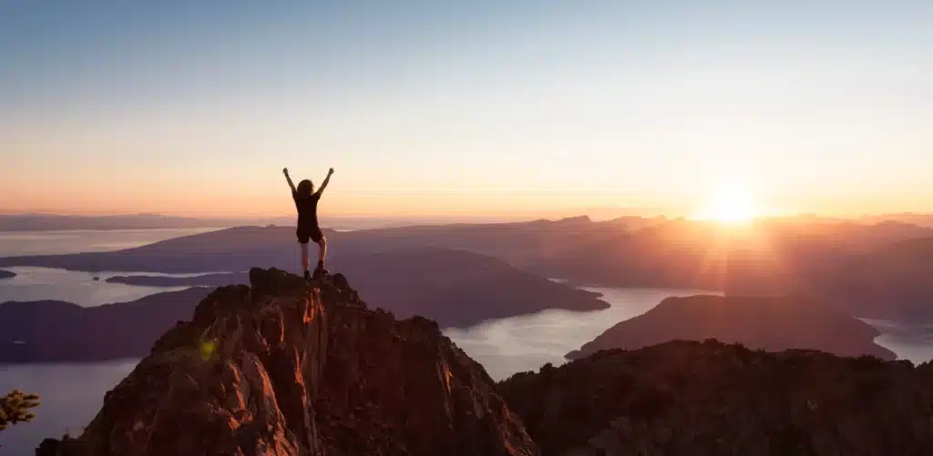 Pessoa celebrando no topo de uma montanha com um pôr do sol ao fundo, destacando uma mensagem motivacional sobre alcançar metas e vencer desafios.